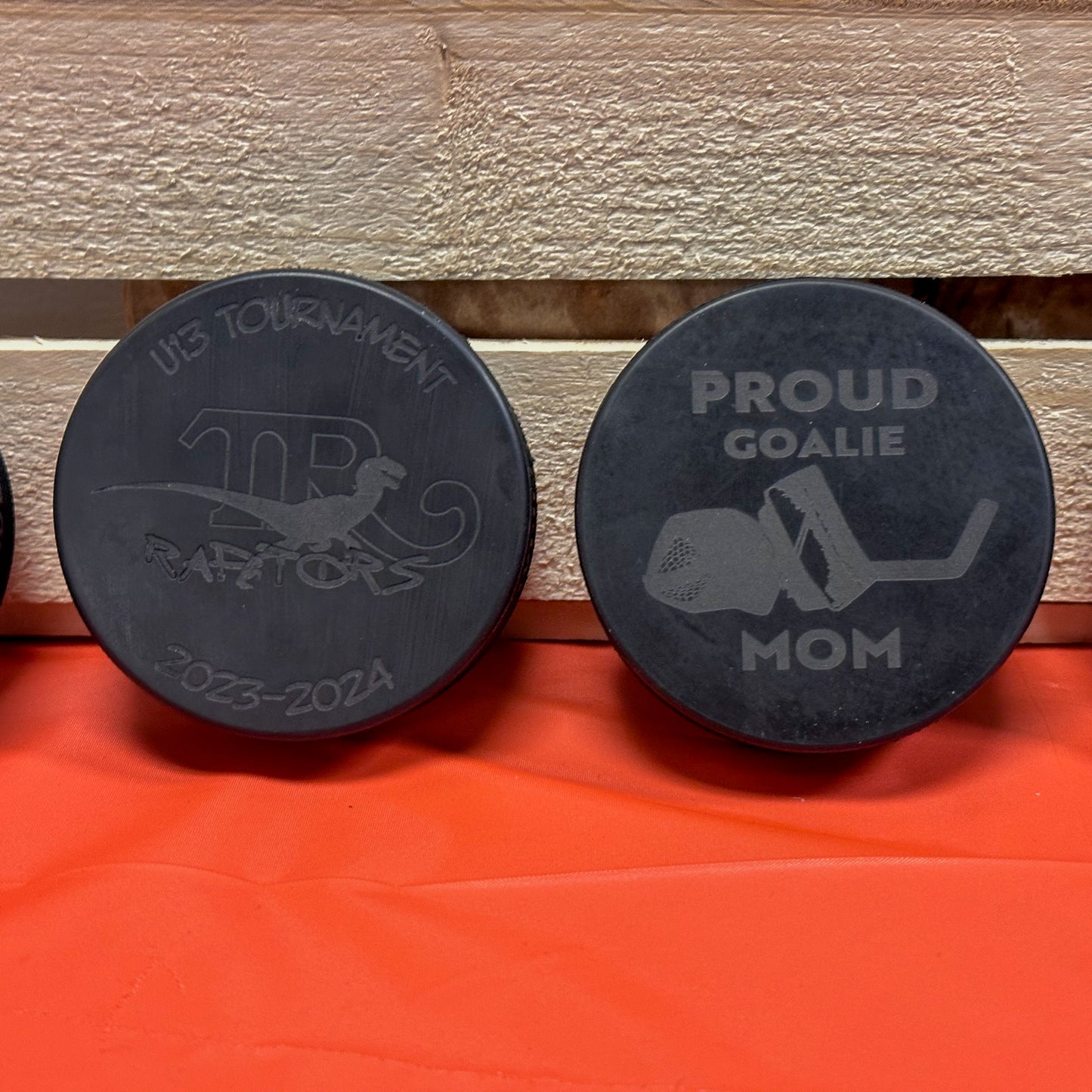 Two engraved hockey pucks rest against a wooden background. The left one is engraved with a team logo and tournament details. The right one is engraved with text reading Proud Goalie Mom with goalie blocker, glove, and stick.