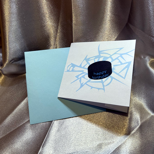 A 4.5" square greeting card sits in front of a blue envelope on a silver backdrop. The card contains an illustration of a hockey puck with the words happy birthday on the side, creating cracks in a light blue surface.