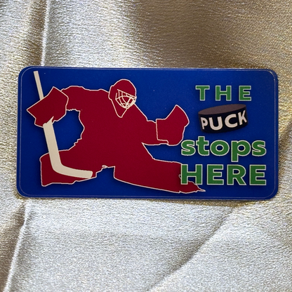 A rectangular sticker with blue background featuring a red silhoutte of a goalie with stick. Text to the right of the goalie reads The puck stops here, with the word puck written on the side of a 3D looking puck.