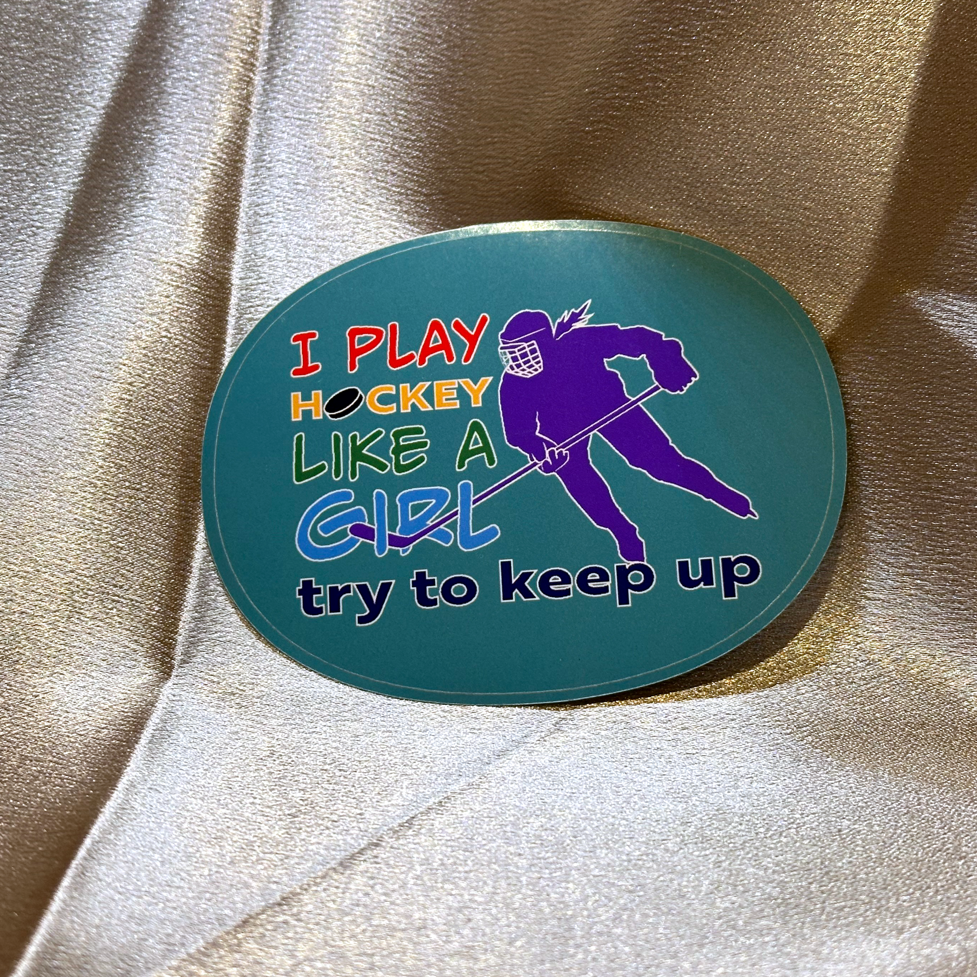An oval sticker with teal background features a purple silhouette of a hockey player with a pony tail over one shoulder. Text in rainbow colours reads I Play Hockey Like A Girl Try To Keep Up.