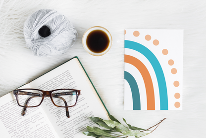 a top-down view of a light colour surface with reading glasses positioned on an open book, a cup of dark liquid, a skien of yarn, and a mockup of a greeting card with a blue and orange rainbow design